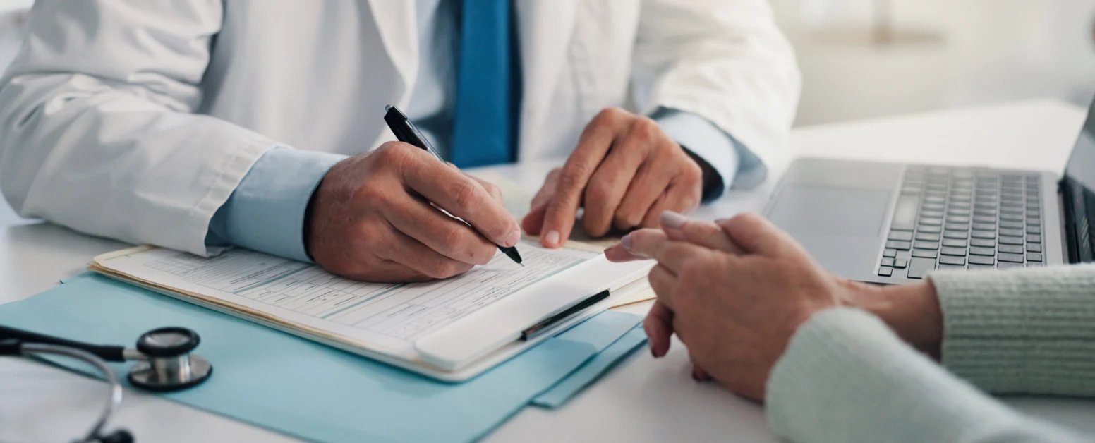 A doctor in a white coat writes on a medical form while talking to a patient across the desk; a stethoscope and laptop are nearby.