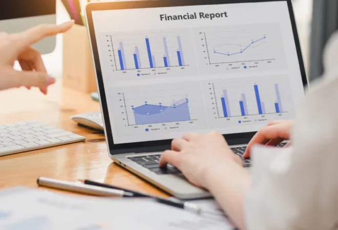 Two people review a laptop displaying a financial report with bar and line charts. One person points at the screen, and documents are spread on the desk.