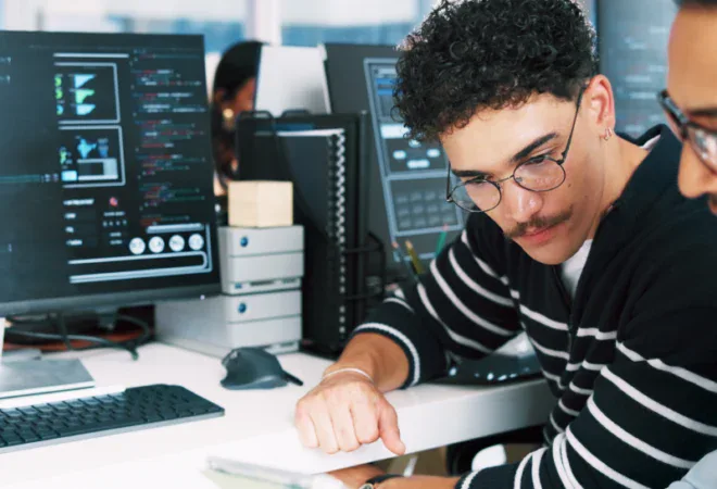 Two people work at desks with computers displaying code; one person explains something on a tablet to the other in an office setting.