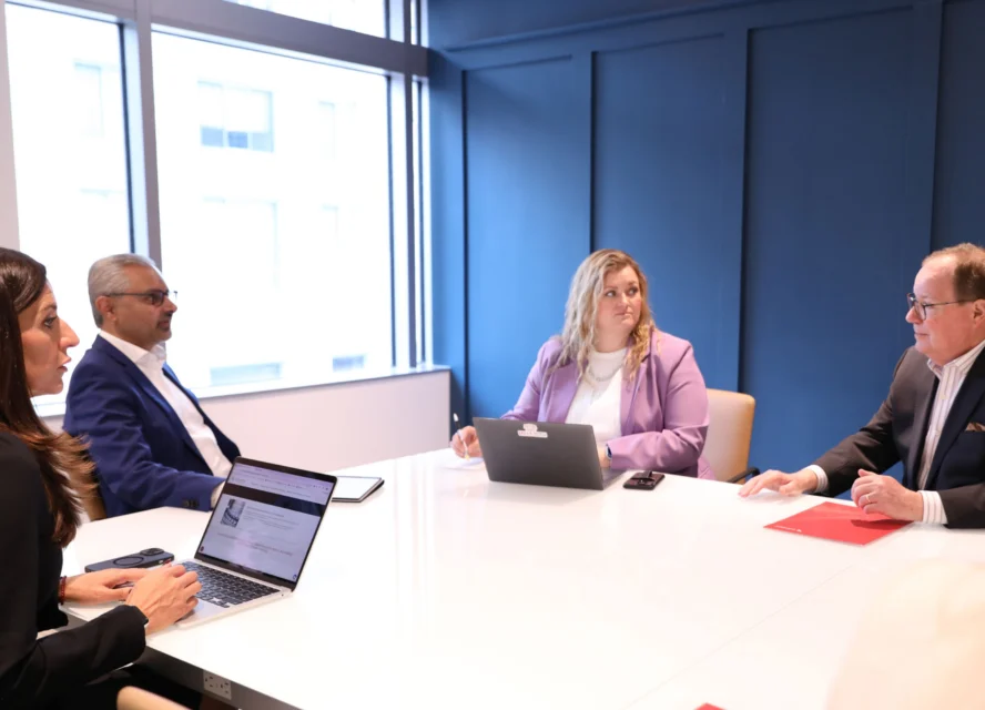 Four people sit around a conference table in a modern office, engaged in a business meeting with laptops and documents.