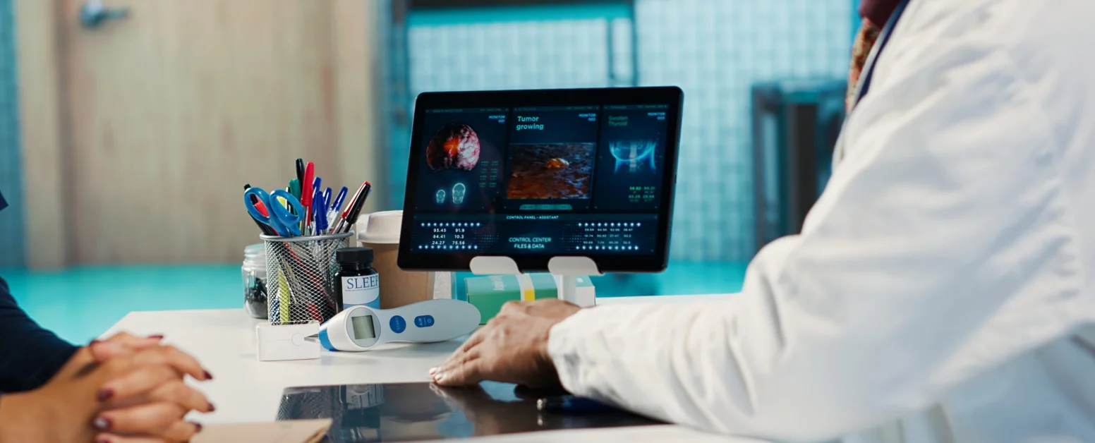 A doctor sits at a desk using a computer displaying medical images and data while talking to a patient whose hands are visible.